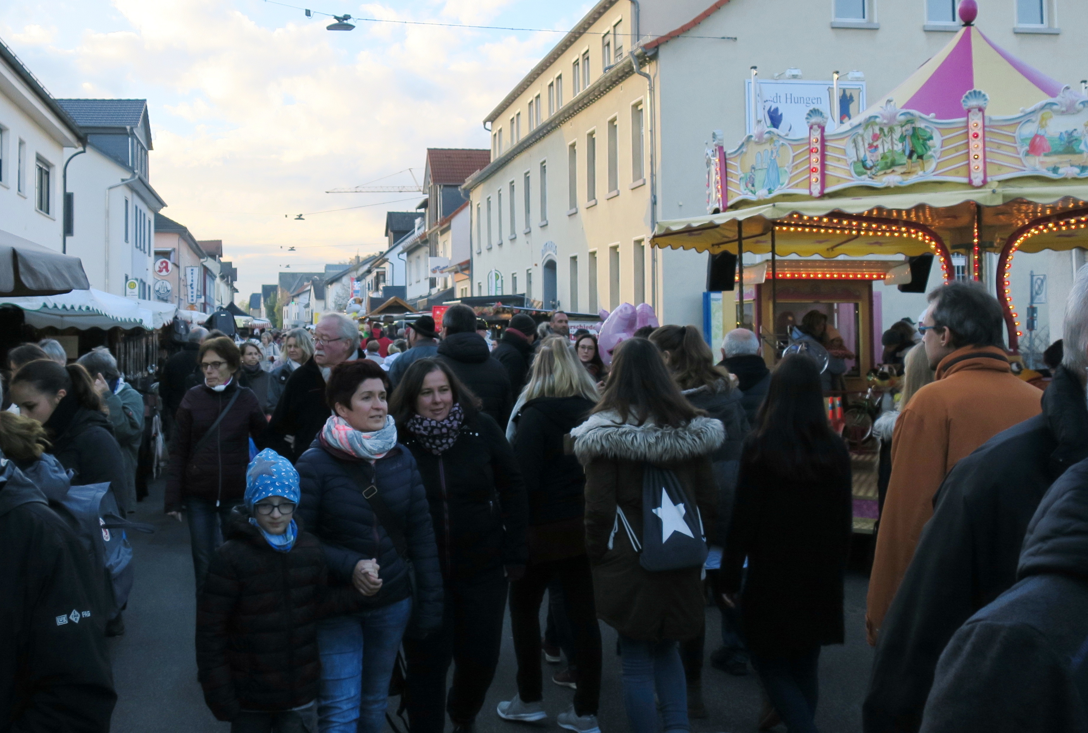 Menschen flanieren durch eine Straße mit Buden an beiden Seiten und einem Karussell rechts. Im Hintergrund das Rathaus von Hungen.