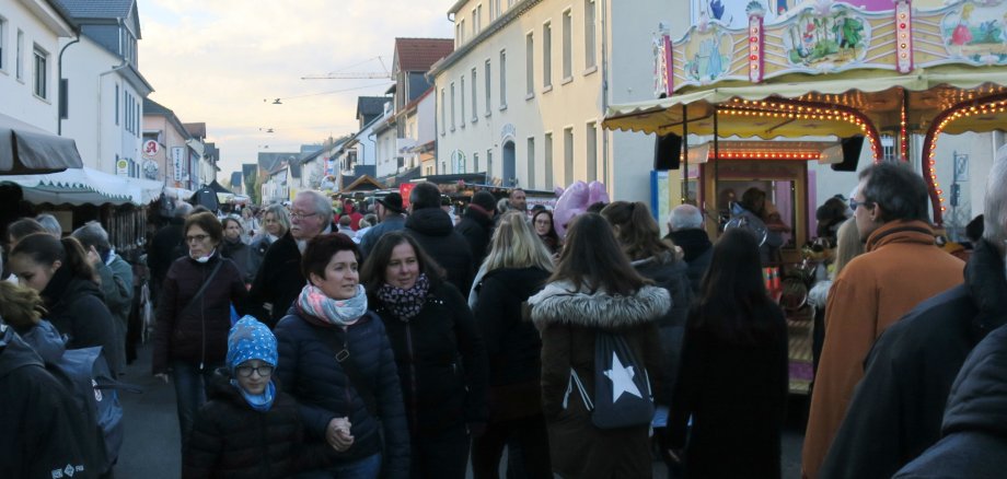 Menschen flanieren durch eine Straße mit Buden an beiden Seiten und einem Karussell rechts. Im Hintergrund das Rathaus von Hungen.