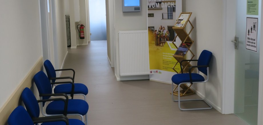 Wartebereich im Flur mit blauen Polsterstühlen, geradeaus rechts zwei Bildschirme, ein Infomationsbanner und ein Prospektständer.