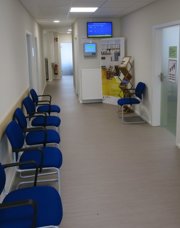 Wartebereich Bürgerbüro Hungen Wartebereich im Flur mit blauen Polsterstühlen, geradeaus rechts zwei Bildschirme, ein Infomationsbanner und ein Prospektständer.