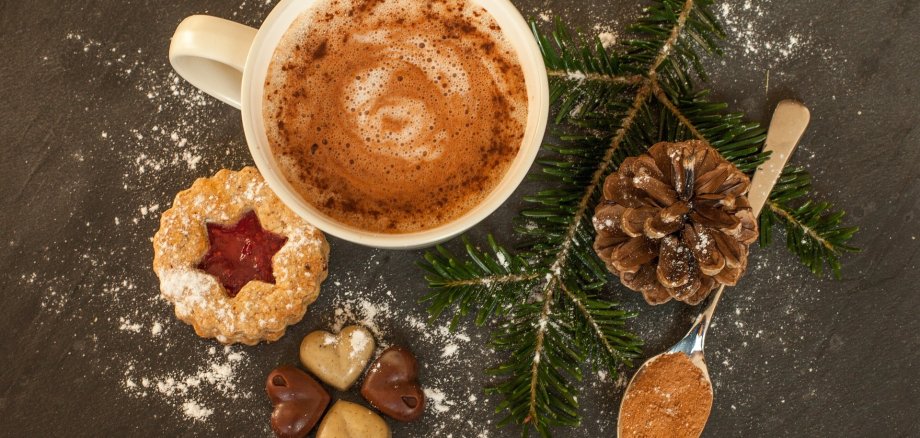 Symbol_Adventsfeier Blick von oben auf eine weißbestäubte Unterlage. Darauf steht eine Tasse Kaffee, daneben liegen von links nach rechts ein Weihnachtskeks mit Konfiturenfüllung, vier Konfektherzen in Form eines Kleeblatts zusammengefügt, ein kleiner Tannenzweig, ein Kiefernzapfen und ein Teelöffel mit Zimtpulver