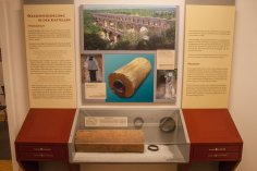 Infokasten zur römischen Wasserleitung Ausstellungskasten mit Teilstück einer römischen Wasserleitung, dahinter Infotafel mit Text und Bildern zur Wasserversorgung in den Kastellen