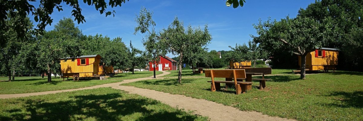 Schäferwagenherberge Nonnenroth: Vier Schäferwagen unter Obstbäumen mit Fußwegen und Servicehaus im Hintergrund, vorne Sitzgruppe