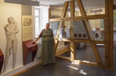 Frau in römischem Gewand vor Rätselstation und Erläuterung zu römischen Bädern